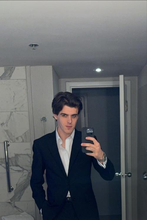 A picture of Callan Stewart standing in front of a mirror in what appears to be his washroom. He is wearing a suit with a black blazer and a white dress shirt.