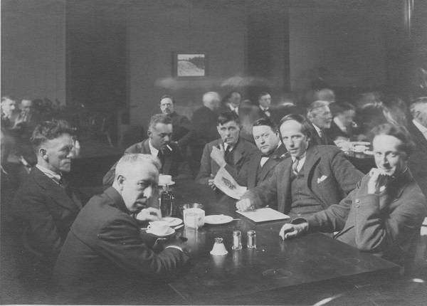 Eight men sitting around a table, with other men at tables in background