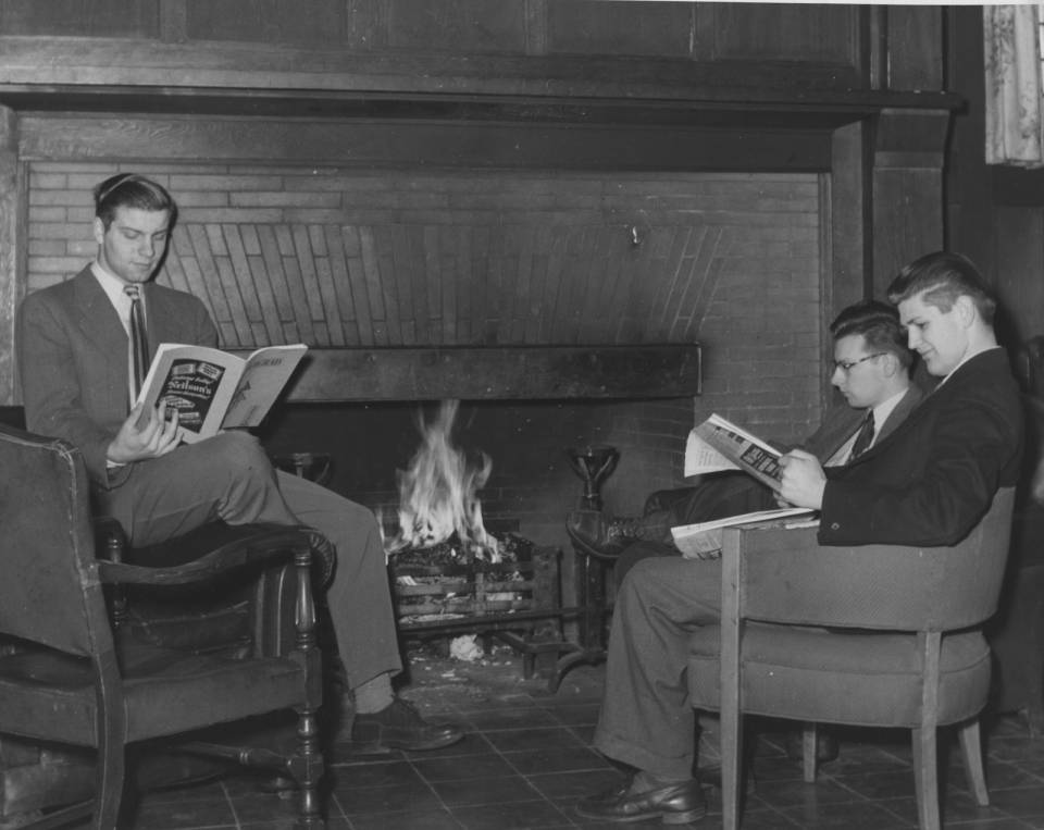 Three young men in suits reading magazines by the fire, in armchairs.