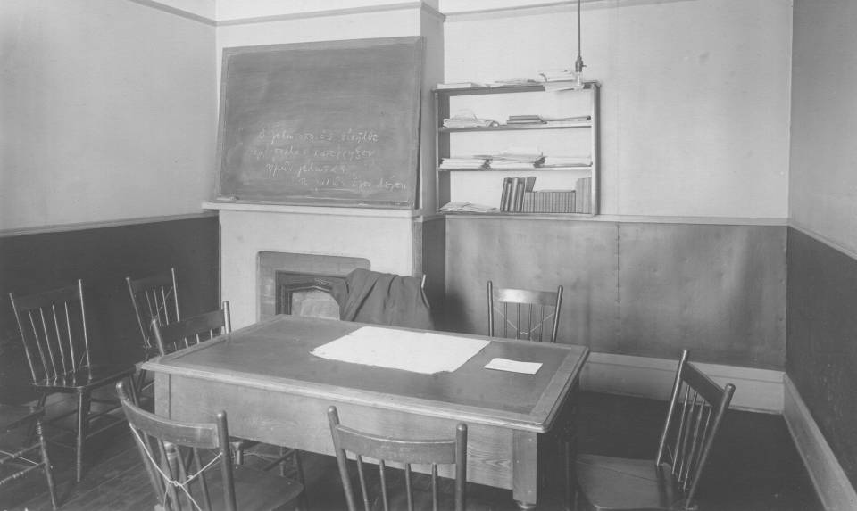 small room containing a table with wooden chairs, a blackboard with Greek writing, and a small bookshelf containing books and papers
