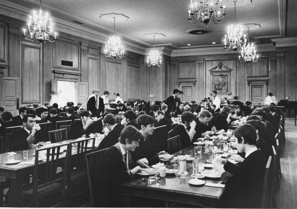 many young men in suits, ties, and academic robes, sitting at long dining tables, eating