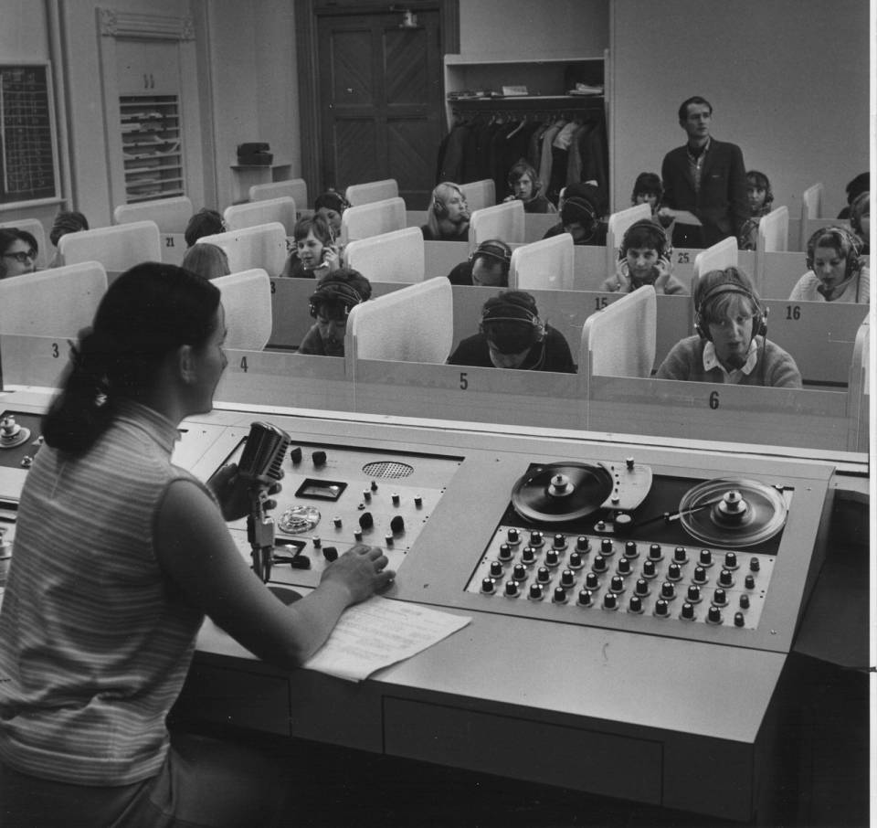 Two teachers and about fifteen students (with headphones) in a language lab