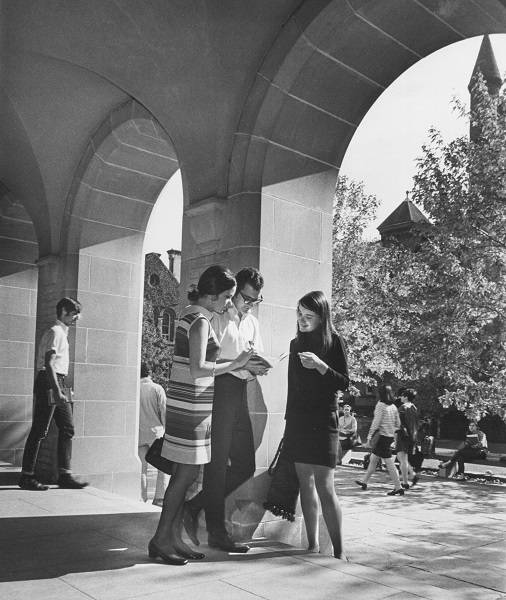 two young women and a young man looking at a book, with University College quadrangle and other students in background