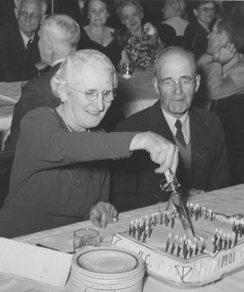 Older woman cutting cake which says "U.C. 1901" while older man looks on