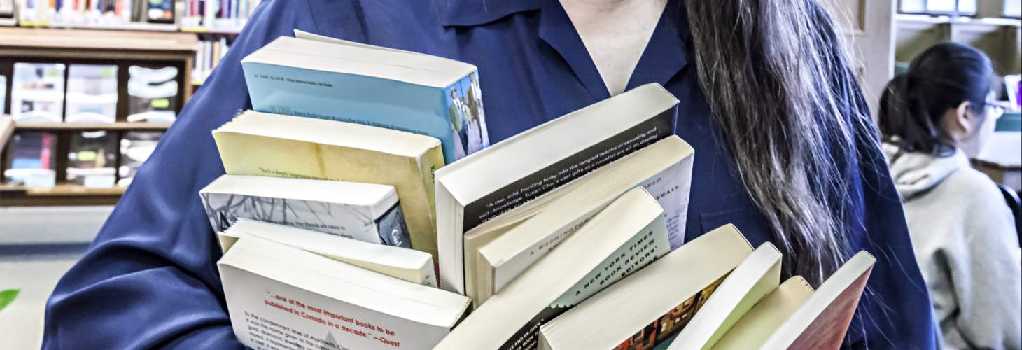 a woman's arms holding twelve books, with library in background