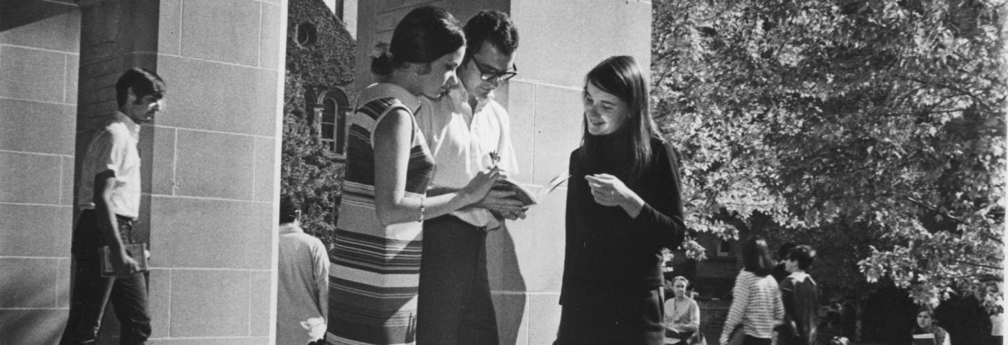 two young women and a young man looking at a book, with University College quadrangle and other students in background