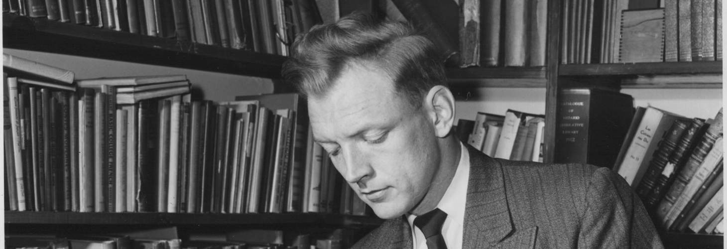 David Hayne, wearing a suit, reading a book in front of a bookcase