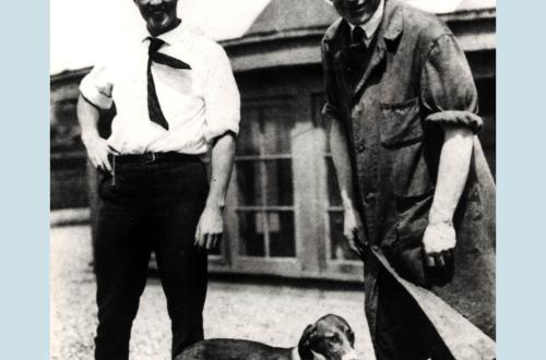 Black & White archival photo of two male scientists standing outside UC with their dog