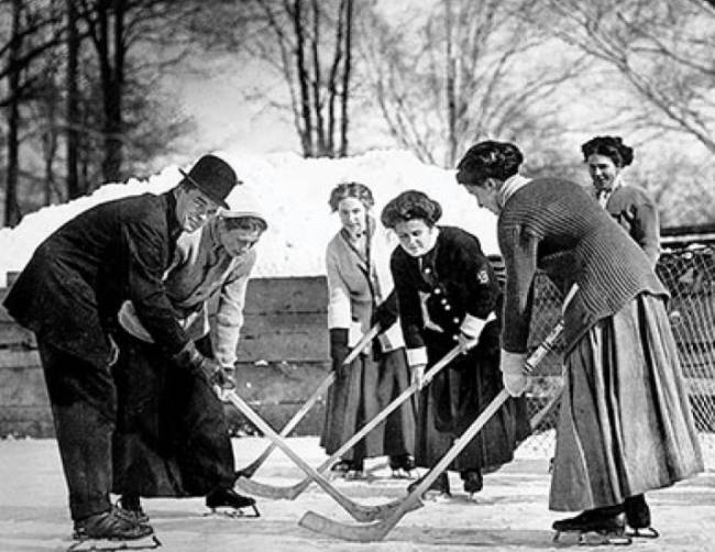 Five women playing ice hockey and a male official holding a hockey puck
