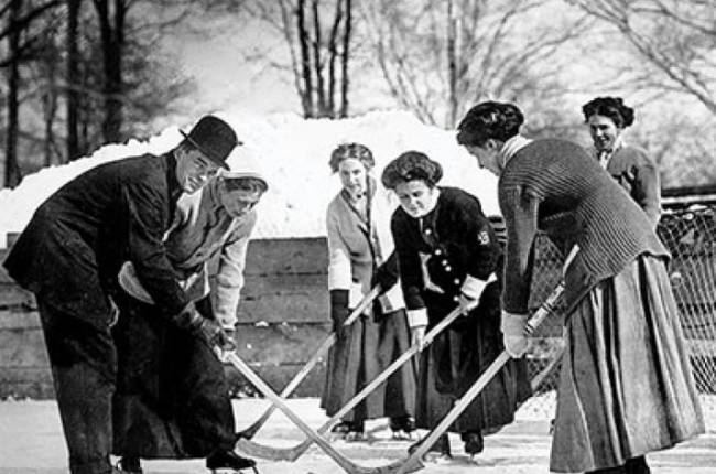 Five women playing ice hockey and a male official holding a hockey puck