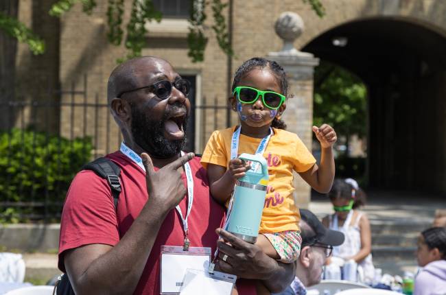 A man smiles broadly as he holds a tiny human (5?). Both are wearing sunglasses and look cool.