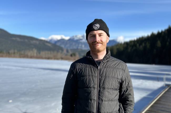 Picture of Liam Fox. The background is scenic and there are mountainous regions. He is wearing a green jacket and black toque.