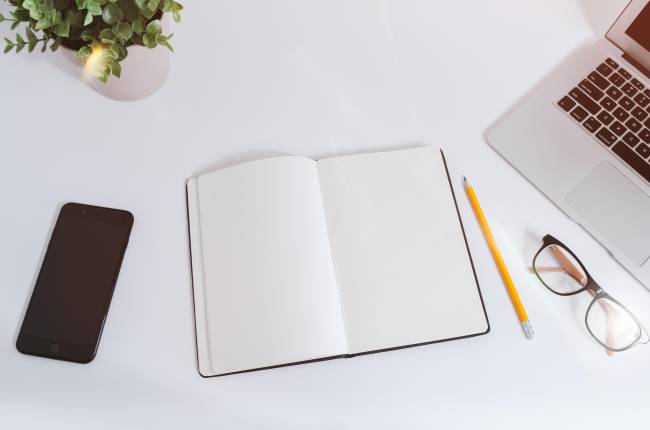 An open notebook on a white desk next to a phone and laptop