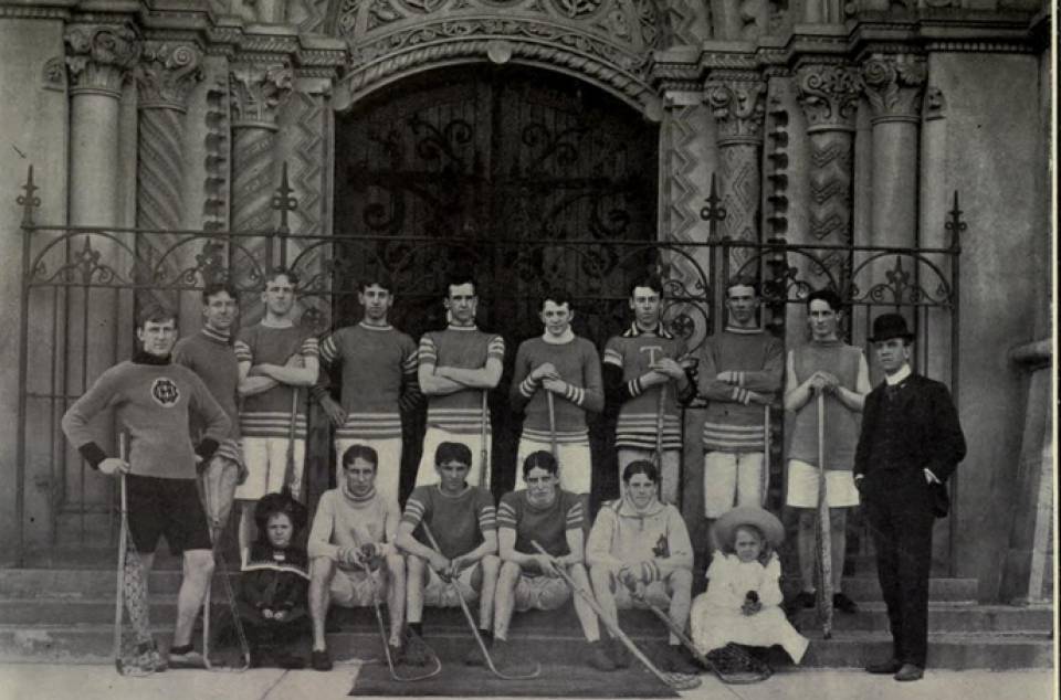 thirteen young men with lacrosse sticks, one man in a suit, and two small children