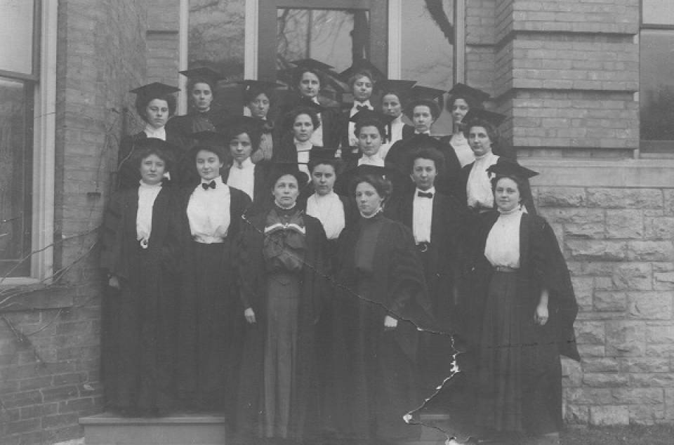 About 20 women posing in doorway of building, wearing academic robes and mortarboards