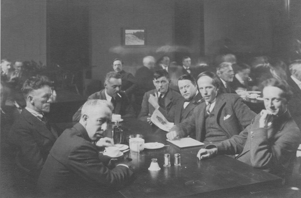 Eight men sitting around a table, with other men at tables in background