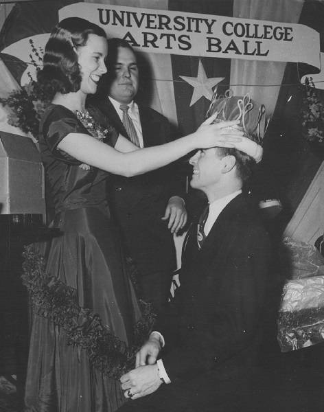 A woman placing a crown on a man's head, with sign in background that says "UNIVERSITY COLLEGE ARTS BALL"