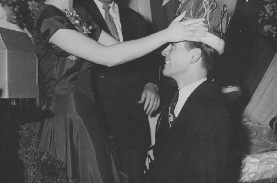 A woman placing a crown on a man's head, with sign in background that says "UNIVERSITY COLLEGE ARTS BALL"