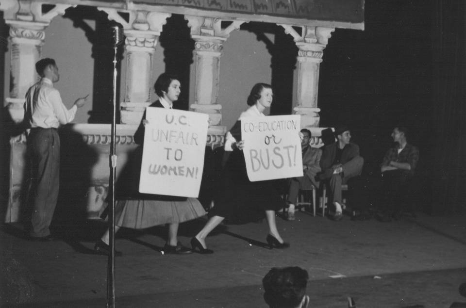 two women on stage holding placards that say "U.C. unfair to women" and "Co-education or bust" with several men in the background.