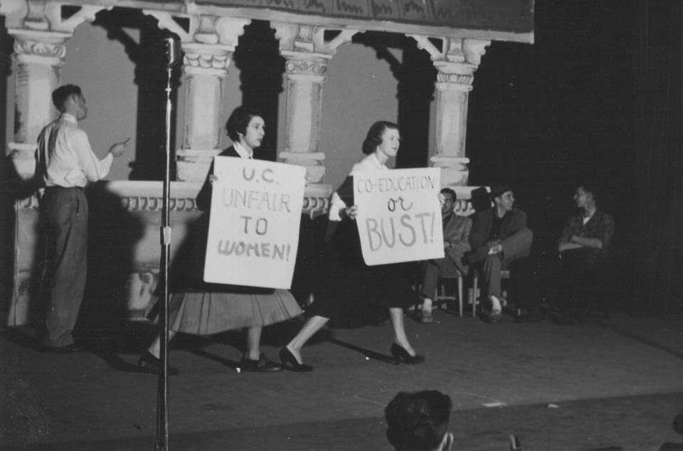 two women on stage holding placards that say "U.C. unfair to women" and "Co-education or bust" with several men in the background.