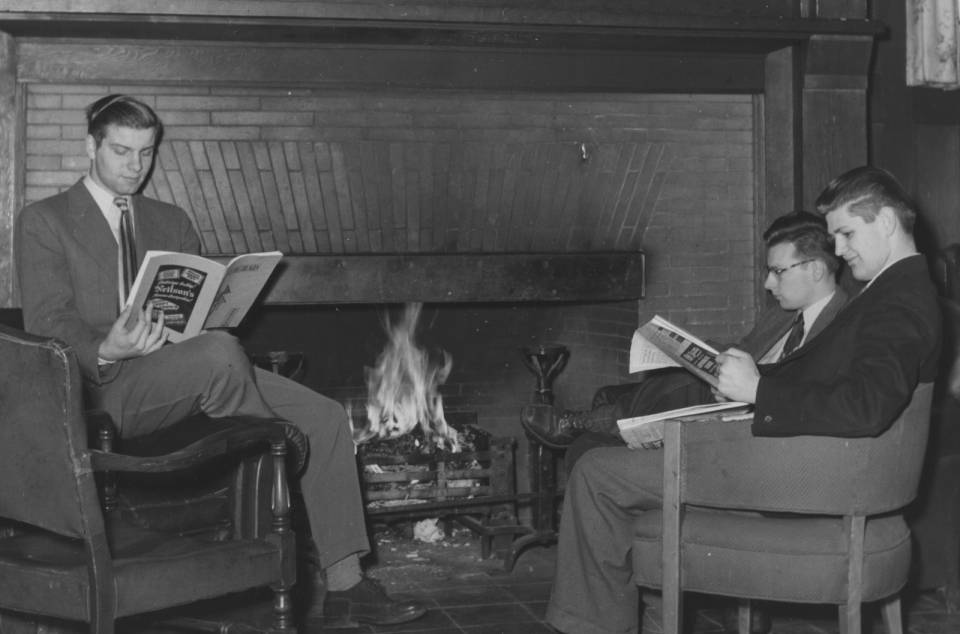 Three young men in suits reading magazines by the fire, in armchairs.