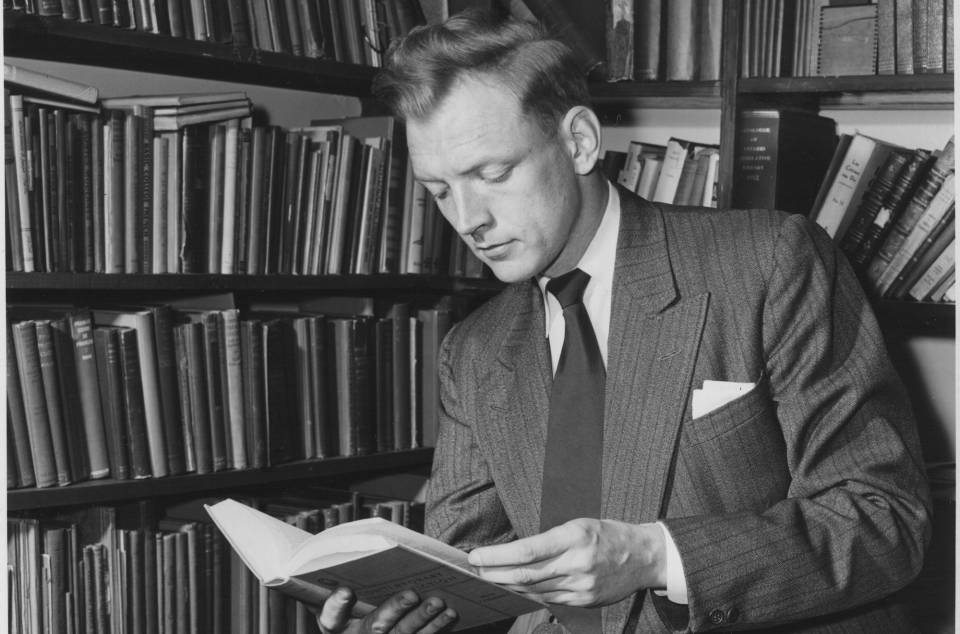 David M. Hayne reading a book, next to shelves filled with books