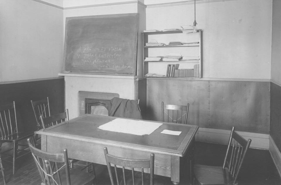 small room containing a table with wooden chairs, a blackboard with Greek writing, and a small bookshelf containing books and papers