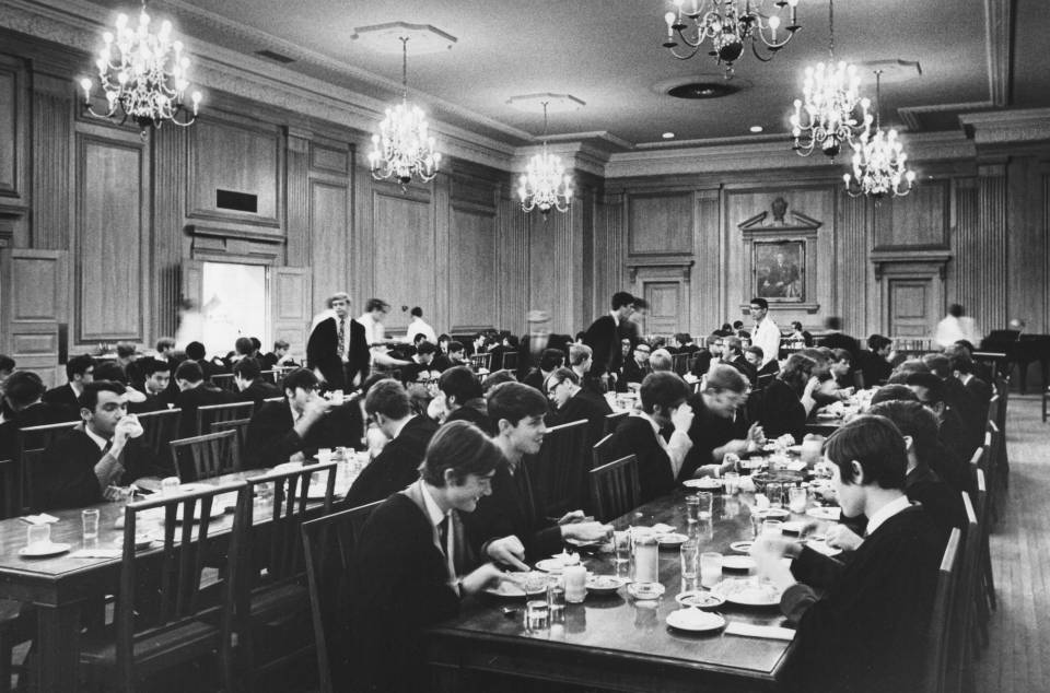 many young men in suits, ties, and academic robes, sitting at long dining tables, eating