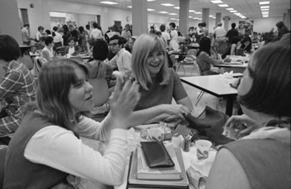 three young women talking at a table, with many other students in the background