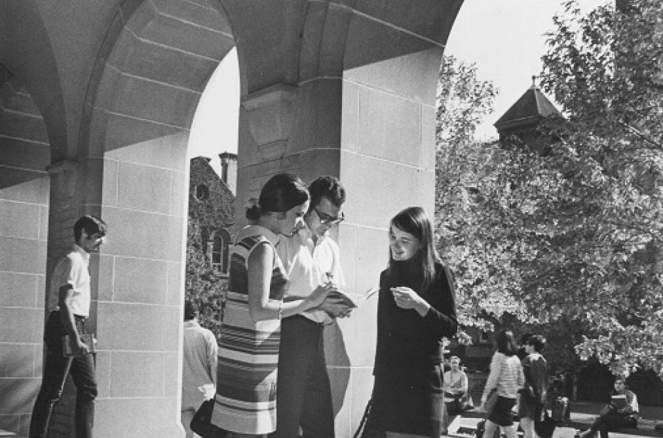 two young women and a young man looking at a book, with University College quadrangle and other students in background