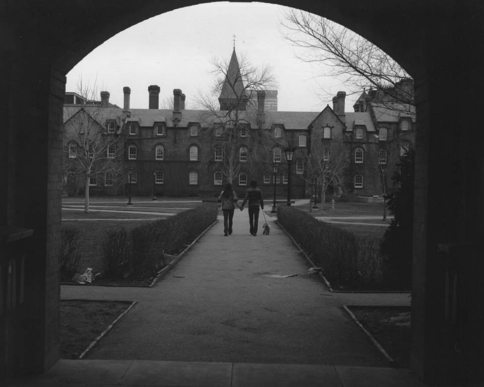 Two people walking with a dog, seen through an archway, with University College in background