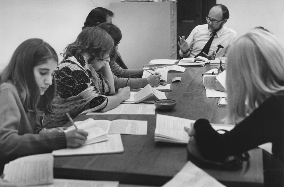 about eight students seated around a table with a professor, taking notes as the professor speaks