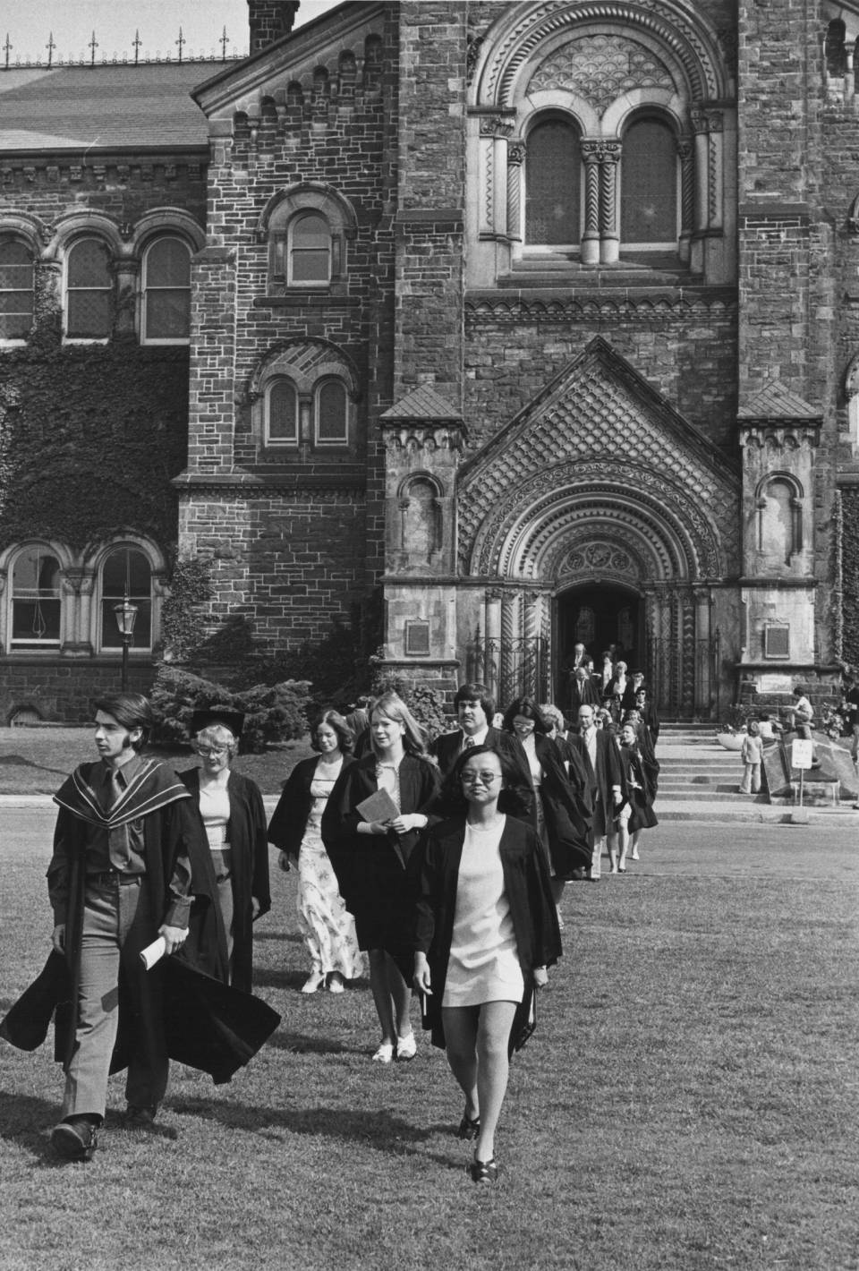 About twenty people in academic robes walking across front campus, with University College in the background