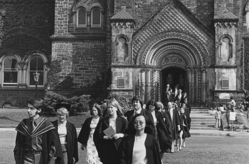 About twenty people in academic robes walking across front campus, with University College in the background