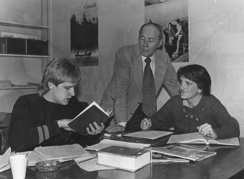 Two students seated at a table with books and papers, and a professor standing next to them