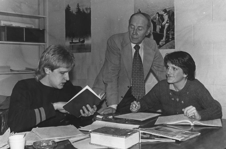 Two students seated at a table with books and papers, and a professor standing next to them