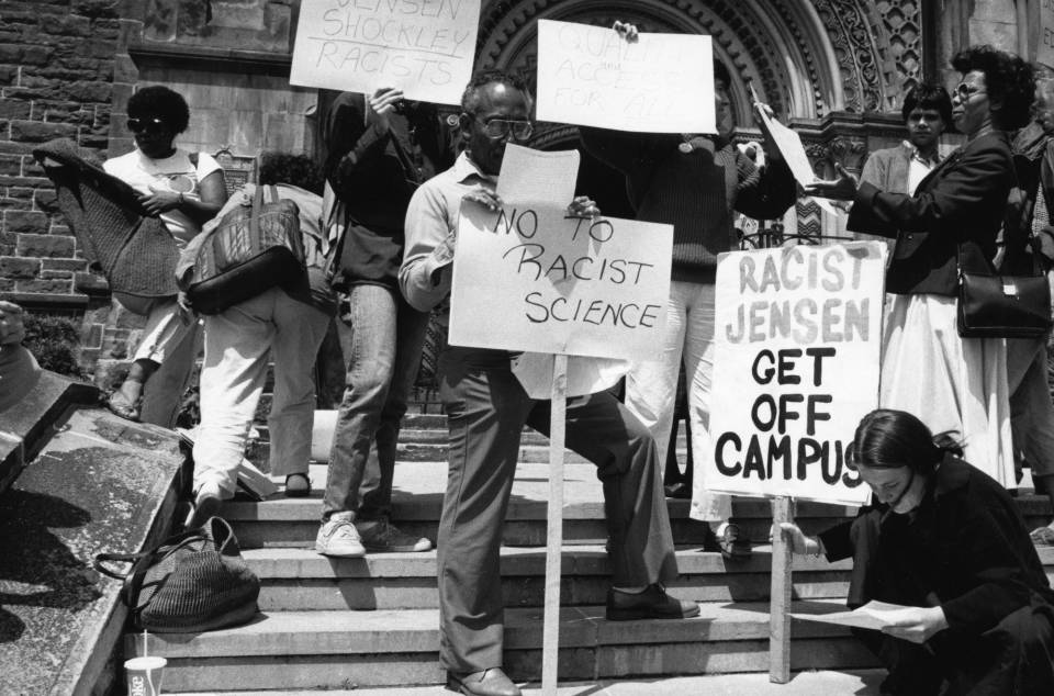 Demonstrators on steps of college, with signs saying "No to racist science" and "Racist Jensen get off campus"