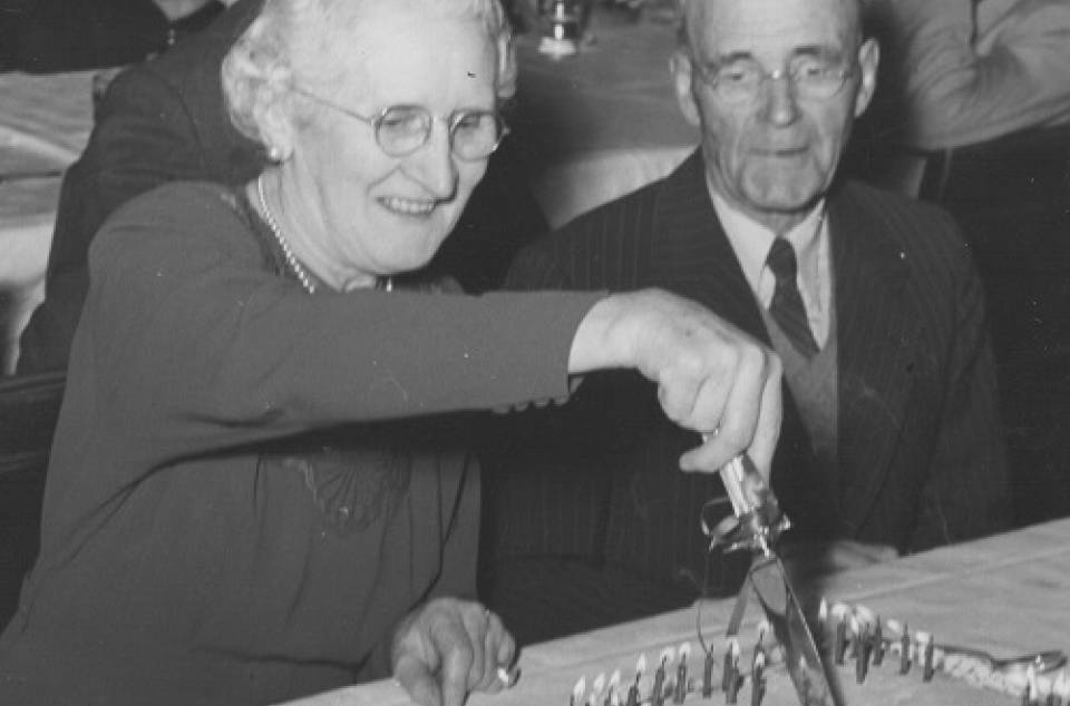 Older woman cutting cake which says "U.C. 1901" while older man looks on