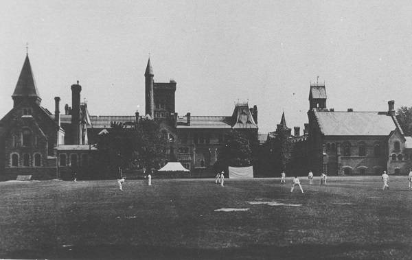 Men dressed in white, playing cricket, with University College in background