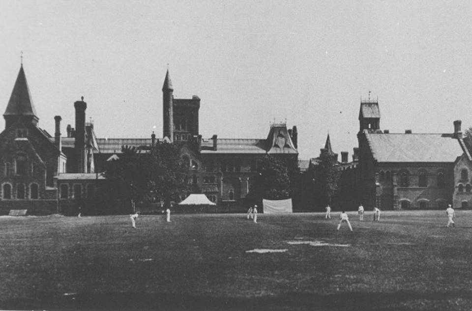 Men dressed in white, playing cricket, with University College in background