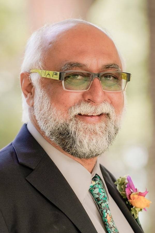 Portrait of Amir Hussain smiling at the camera, wearing a dark grey suit, pale grey shirt and bright blue and green impressionist floral tie with an orange and pink boutonnière in his lapel. H