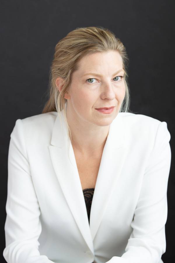 Portrait of Maev Beatty, a woman with blond hair pulled gently back and wearing a natural look in a crisp white blazer with a black shirt barely visible. Her face is turned slightly to the left but she is looking at the camera with a slight smile.