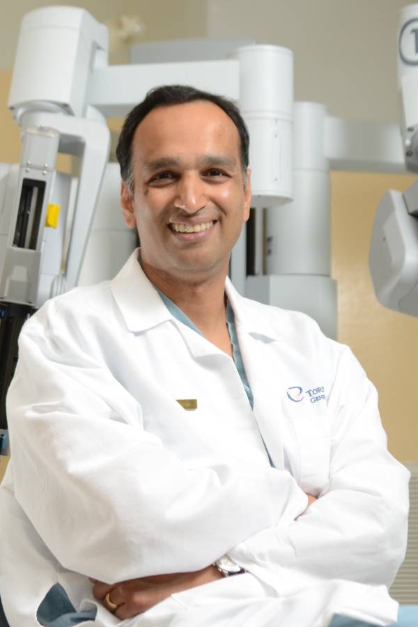 Dr. Singal wears a white lab coat, seated in front of a Da Vinci Surgical System machine. He is smiling at the camera with his arms folded in his lap.