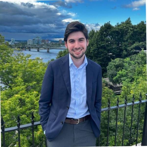 A picture of Jonah Rosen. In this image, Jonah is resting on a metal fence with his hands tucked in his pockets. There are trees in the background.