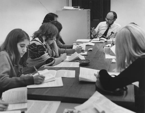 about eight students seated around a table with a professor, taking notes as the professor speaks