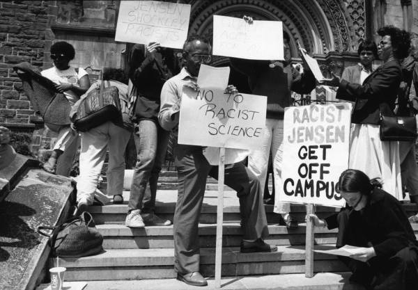 Demonstrators on steps of college, with signs saying "No to racist science" and "Racist Jensen get off campus"