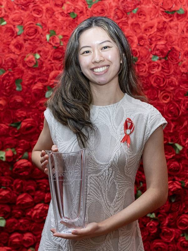 Diana Lam stands holding her AOI award - a glass vase - smiling for the camera. A red ribbon with a UC pin is pinned to her offwhite dress.