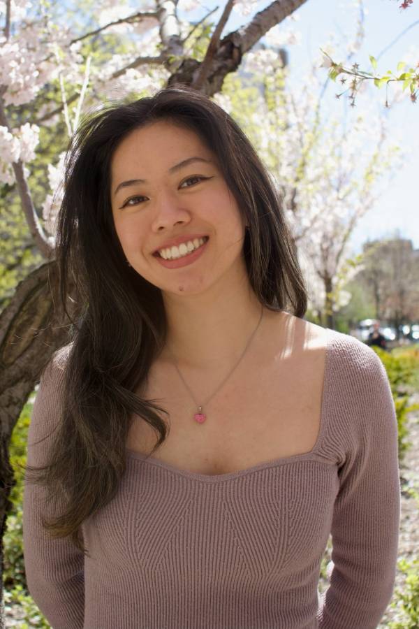 Outdoor portrait of Diana Lam, her head tilted slightly to the right as she smiles at the camera. in a beige rose shirt with her long black hair falling over her right shoulder. The out-of-focus tree behind her appears to be in bloom.
