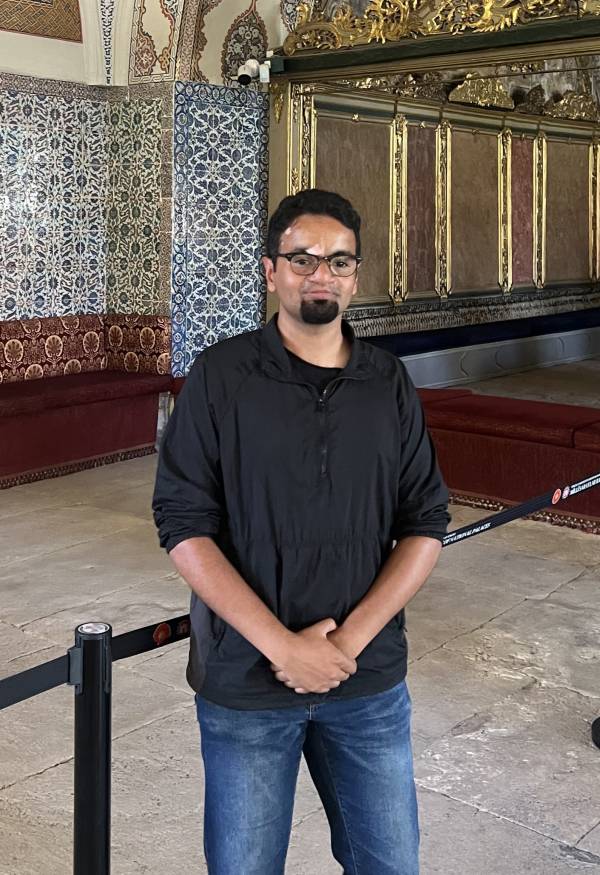 A picture of Talha Hasnat, UC student, posing in a building with intricate designs on its walls. Talha is wearing a black t-shirt with a black flannel, and blue jeans. He is smiling. 
