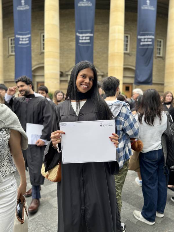 Kiara Andrade wears academic regalia and holds degree in hand while standing in front of columns